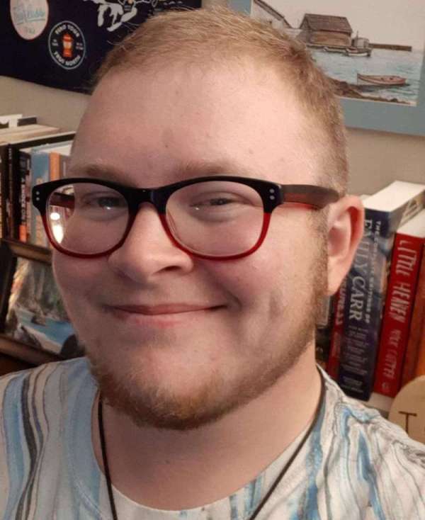 Headshot of Lys, a white man wearing red and black rimmed glasses. He is looking at the camera and smiling. There is a row of books behind him.