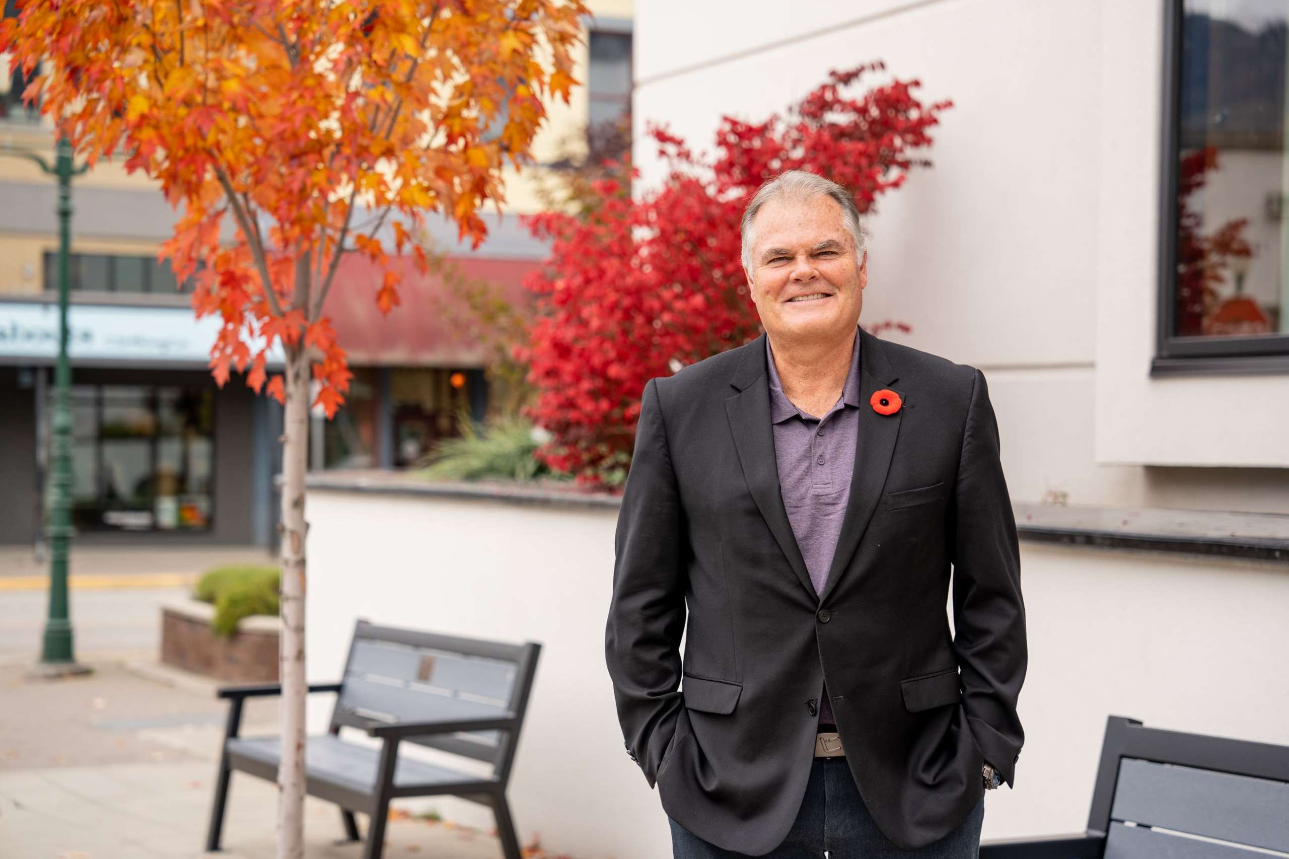 Evan Parliament, an older Caucasian male in a suit standing in front of city hall with a poppy pinned to his suit jacket. He is smiling at the camera with his hands in his pockets looking relaxed.