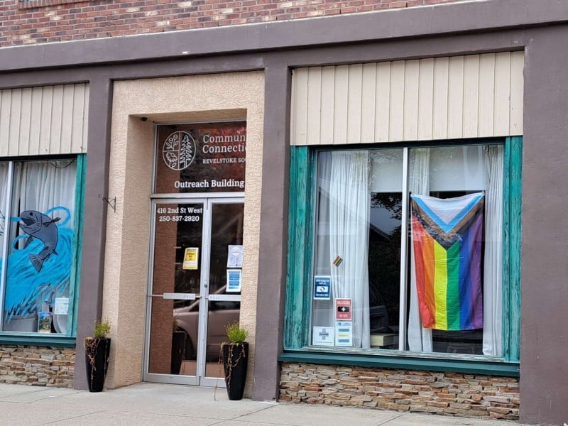 The front entrance to the Revelstoke Community Connections outreach building. In the left hand window is a mural of fish in a river. Handing in the right hand window is an updated Pride flag with the BIPOC and Trans Flag colours and the Intersex symbol.