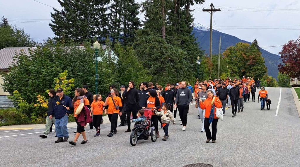 Marchers for orange shirt day come down a sloped street. The angle of the slope allows you to see the whole procession of marchers.