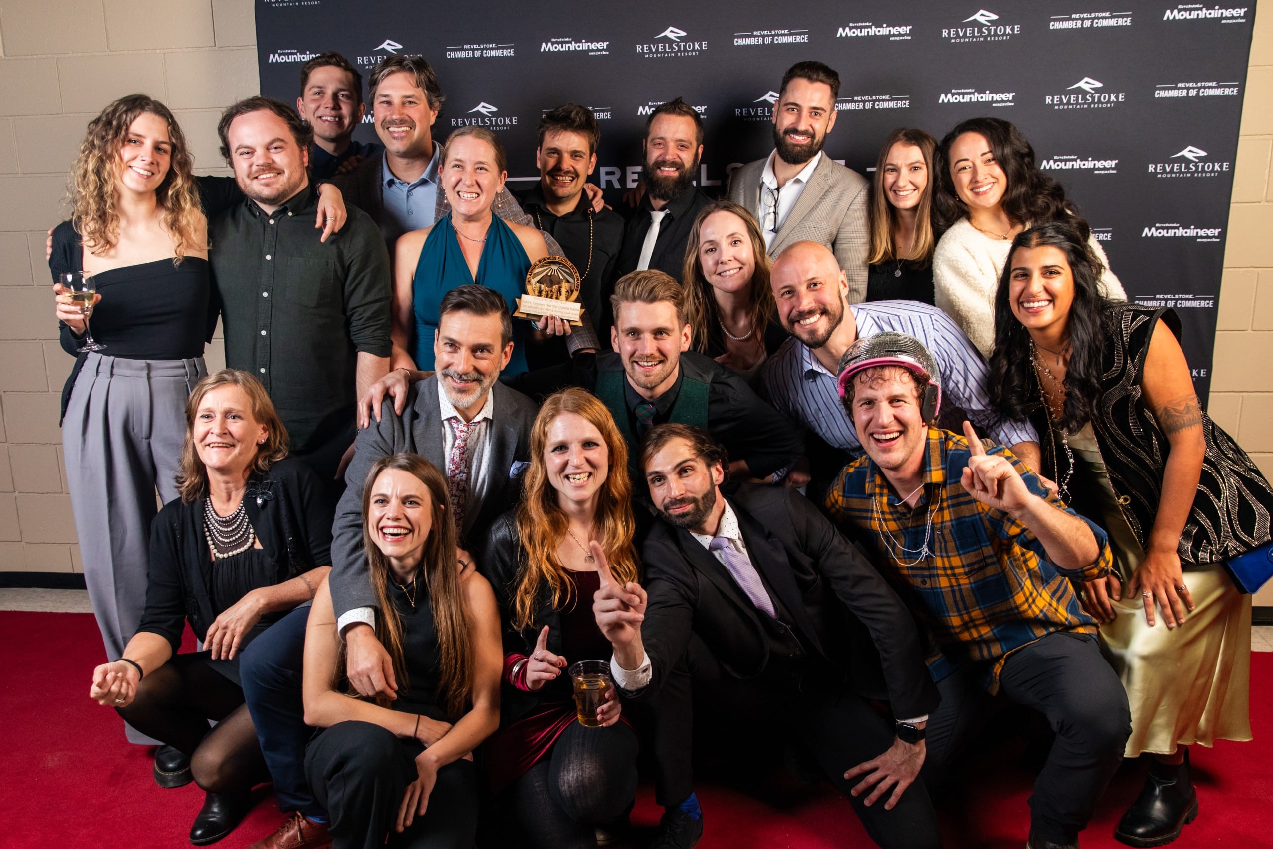 A collection of 20 individuals in business formal wear stand in front of a Revelstoke Business Excellence Awards banner, all smiling at the camera.