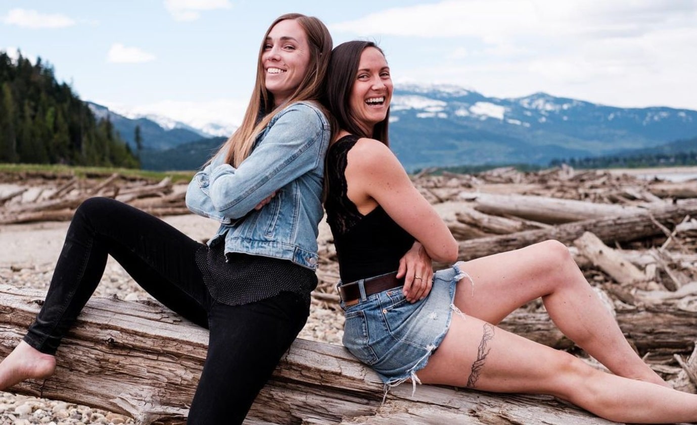 THE STUDIO owners Jacqui Gardiner and Kenley Knock sit back to back on a log, arms crossed staring at the camera with a smile. Both are younger white women with dark brown hair longer than shoulder length,.