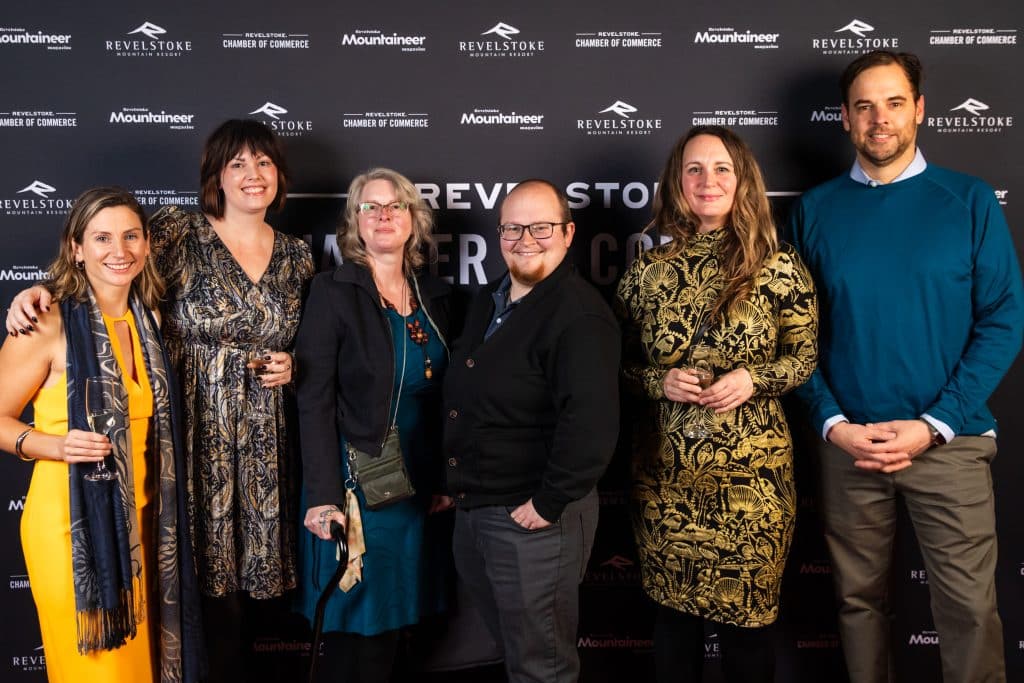 Six people in business formal dress stand in front of a Revelstoke Chamber of Commerce banner. They are all looking at the camera, side by side, smiling.