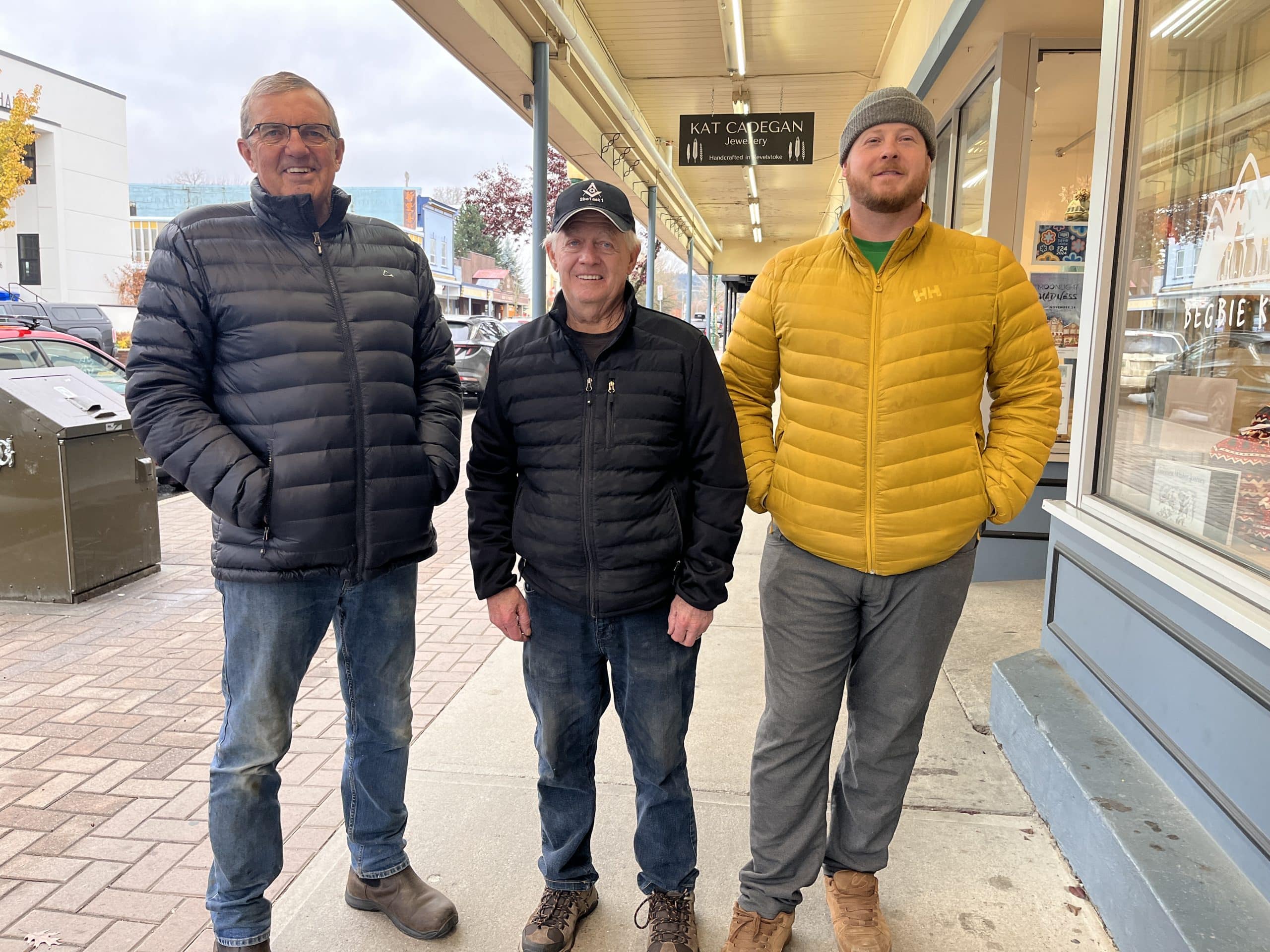 Three who are a part of the Revelstoke Masons stand outside. The one on the left is tall and in a black coat. The middle man is shorter and in a black coat. The man on the far right is tall and ina. yellow coat.