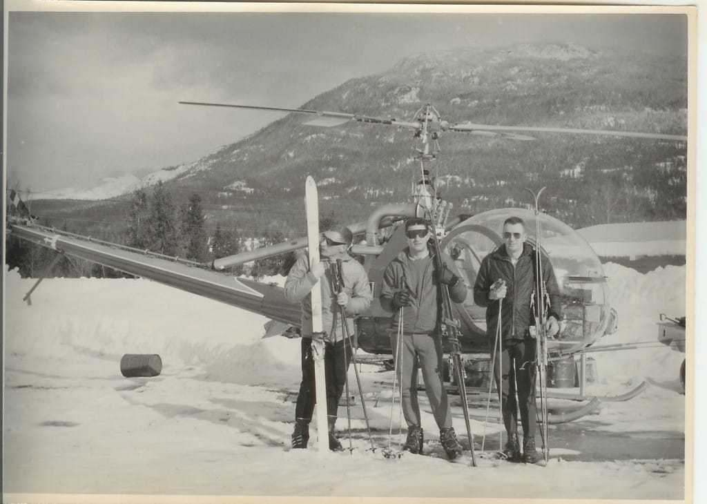 Three men stand on a mountain in front of a helicopter with skis in a clearly dated photo in black and white sepia tones