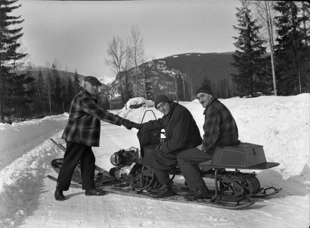 One man stands beside a snowmobile on a snowed road. Two other men sit on the snowmobile. A mountain is in the back ground and the photo is black and white.