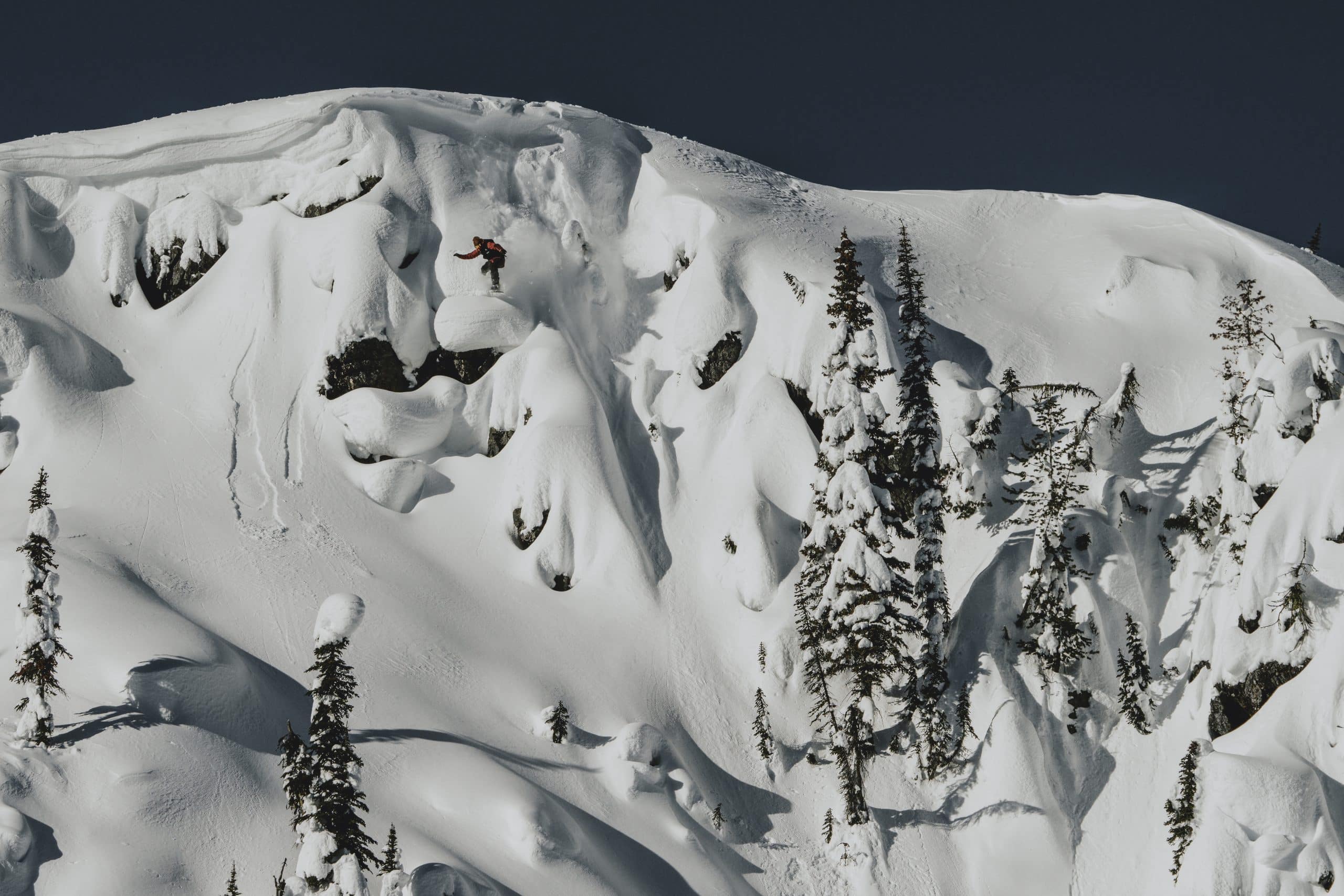 Dustin Craven on a snowboard jumping from a rock face covered with thick snow.