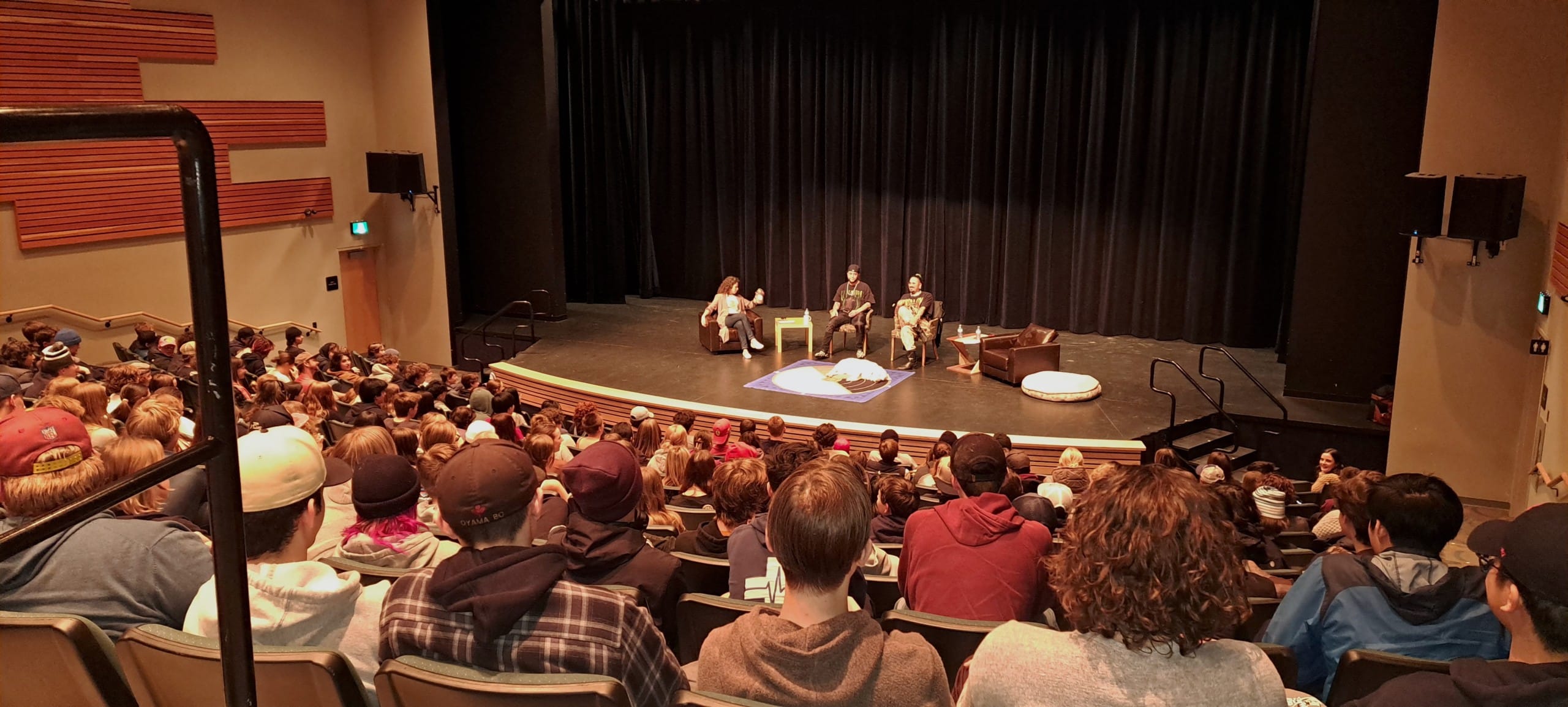 Photo looks down at a stage where rappers Snotty Nose Rez Kids sit with a speaker. In front of them are rows and rows of students, the image is behind them and showing all the backs of their heads.