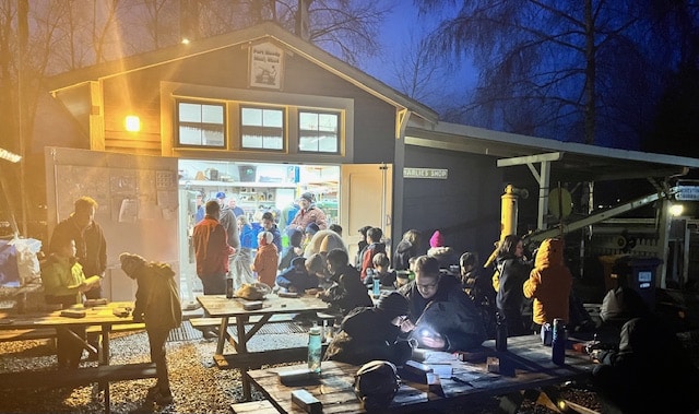 A night picture of a Men's Shed in Port Moody where a number of men are standing around picnic tables in the snow outside of a well lit building. 