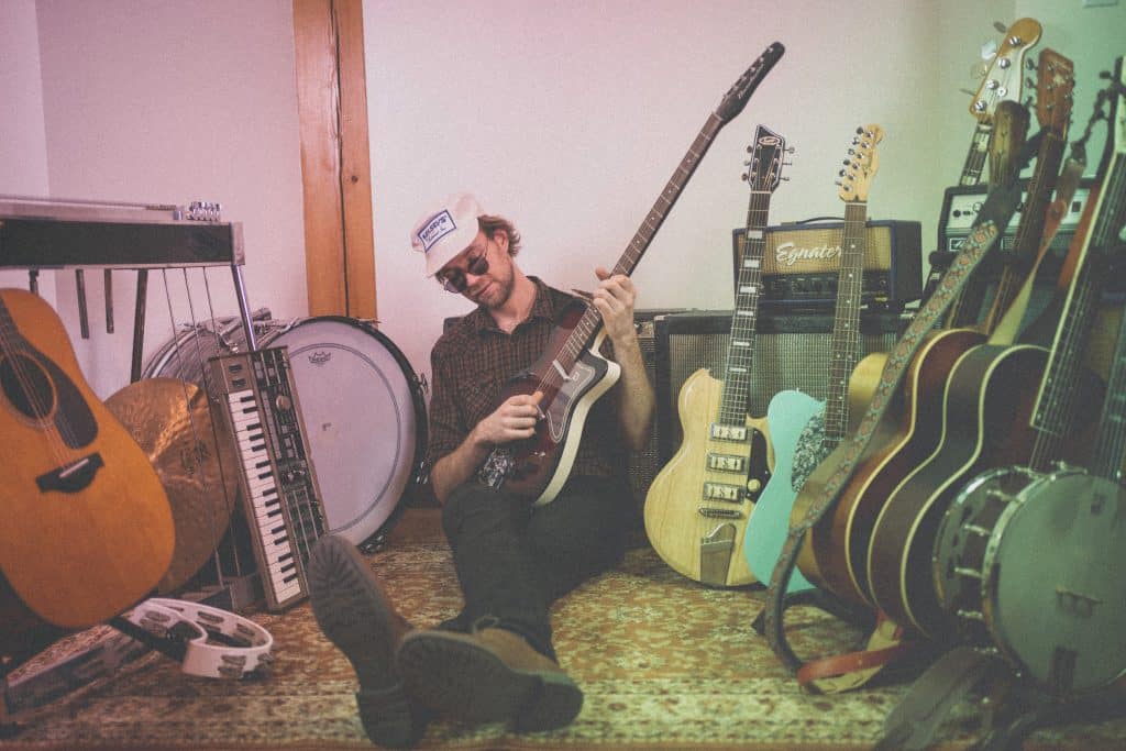 Porch Hounds’ Nik Winnitowy sitting on the floor with his back against an amp. He is playing one electric guitar with a lineup of guitars flanking either side of him.