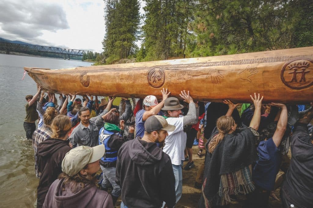A crowd of over 20 people lift a large wooden canoe out of the water and up the banks of a river. The canoe is carved with hand prints, figures holding hands and representation of the land.