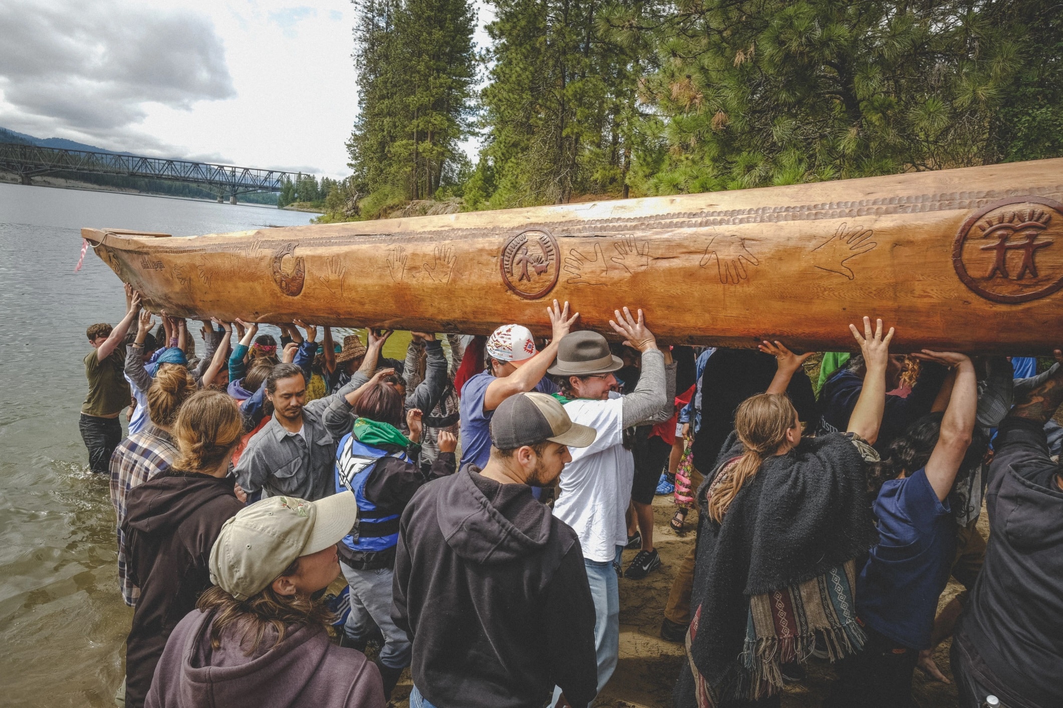 A crowd of over 20 people lift a large wooden canoe out of the water and up the banks of a river. The canoe is carved with hand prints, figures holding hands and representation of the land.