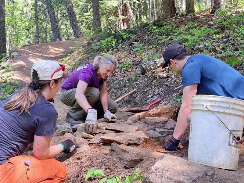 Dig Day volunteers all crouching around a chunk of mountain bike trail, adding stones and dirt to create a path over a large tree root.