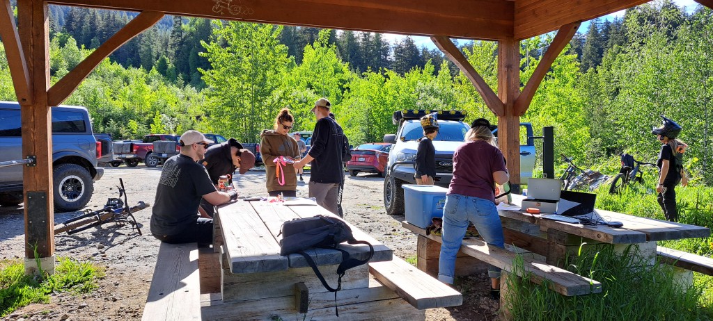 Revelstoke Cycling Association volunteers gather around a picnic table under a gazebo. Bikers stand outside the gazebo in the sun waiting to register.