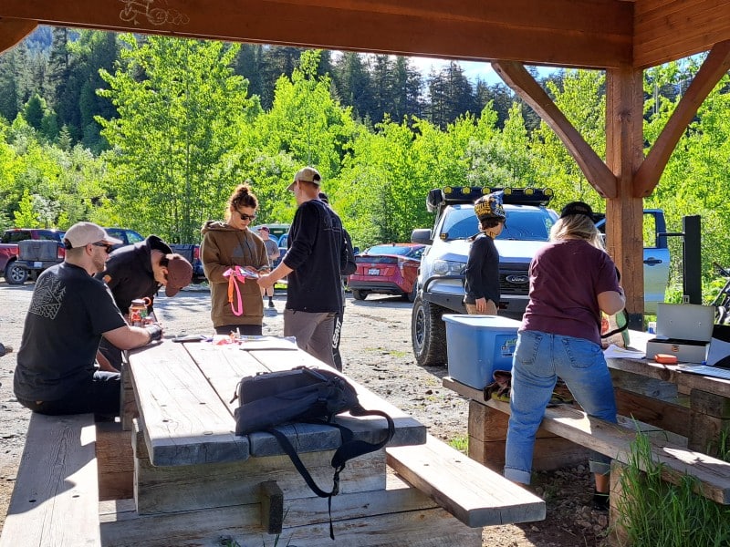 Revelstoke Cycling Association volunteers gather around a picnic table under a gazebo. Bikers stand outside the gazebo in the sun waiting to register.