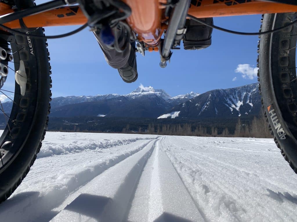 Accessibly cycling route on a snowy flat trail outside Revelstoke. Shot taken from the underside of a recombant bike looking through the front wheels onto the snow flats with trees and mountains in the distance.