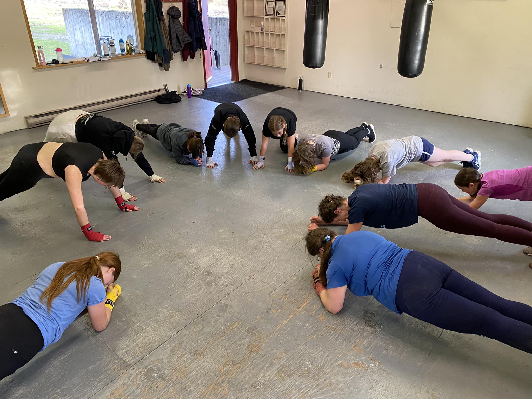 Revelstoke boxing club coach in a plank form with a circle of Girl Guides youth also in various stages of plank formation. There is a hanging heavy bag and some extra boxing gloves along a wall in the back.
