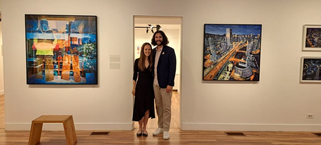 Two figures standing side by side in a doorway with two large paintings on the walls on either side of the door.