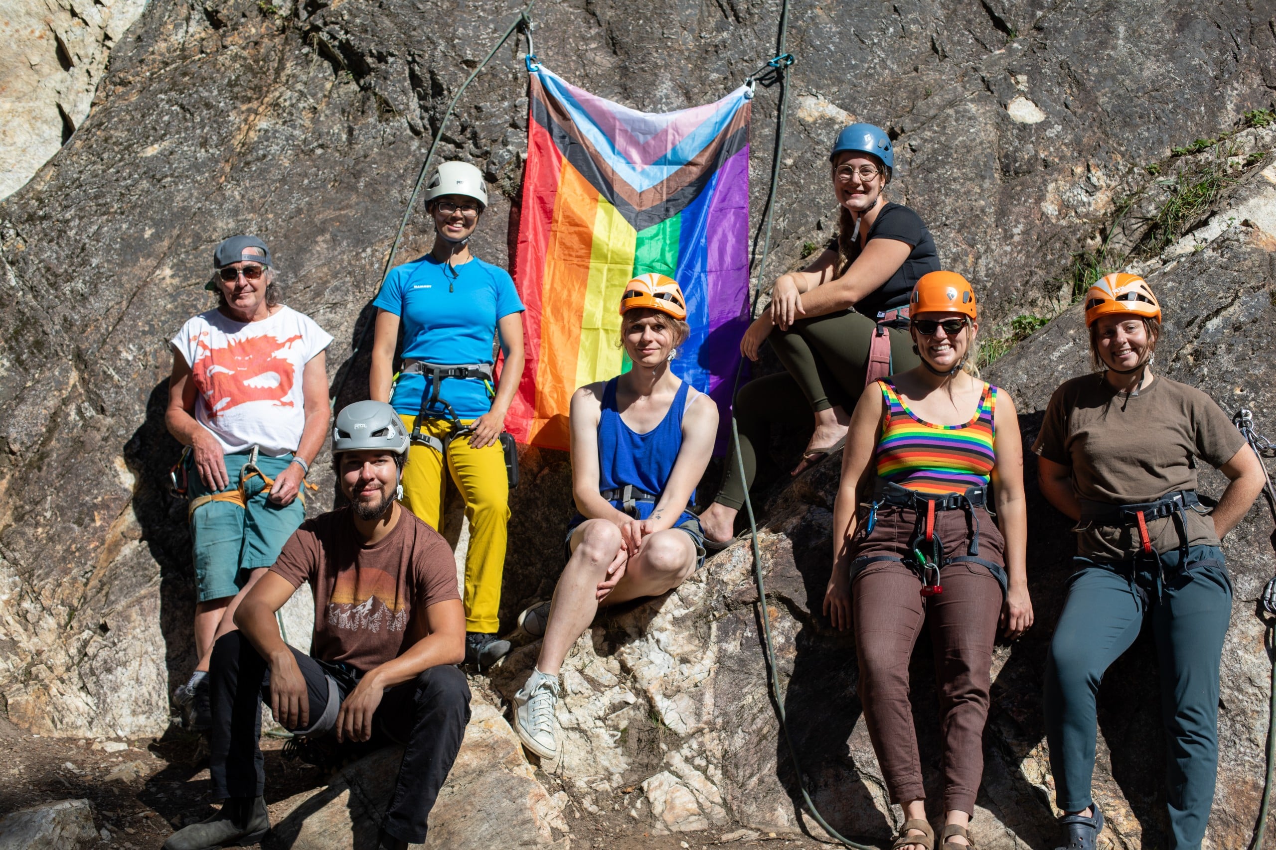 A group of seven individuals, all in rock climbing gear, sit and stand against a rock face and smile at the camera. Behind them is the inclusive pride flag with the intersex triangle added.