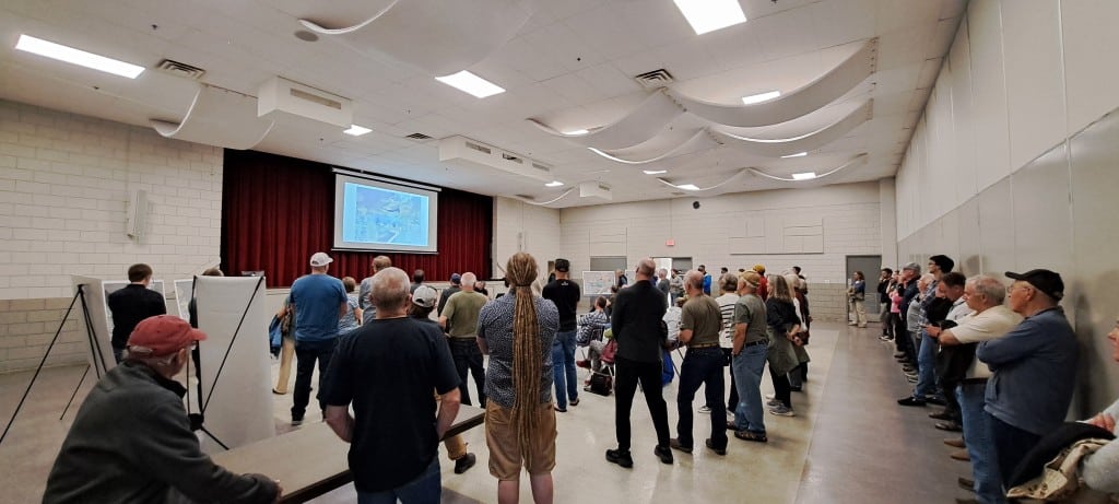 Revelstoke Mountain Resort June open house participants stand around in a large room staring at a screen and two presenters at the front.