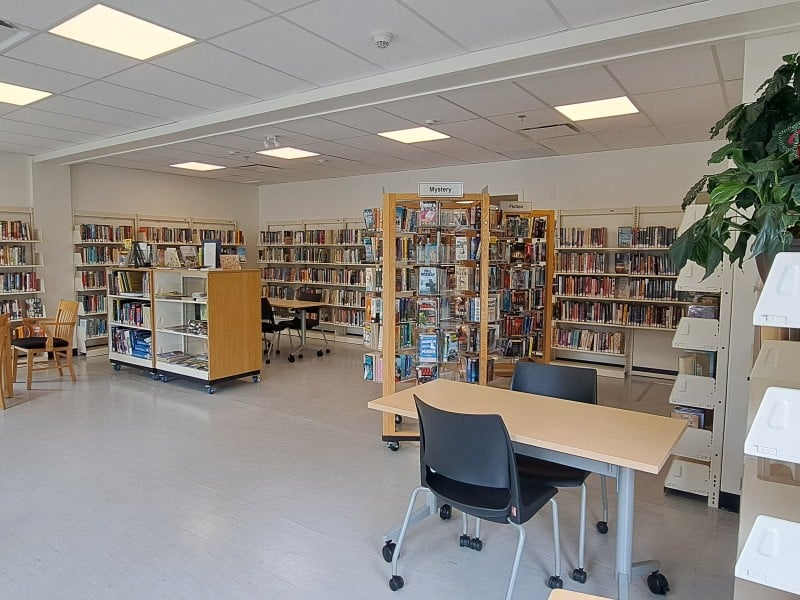 Revelstoke library new space with multiple shelves of books, white tile, a collection of desks and chairs and large windows.