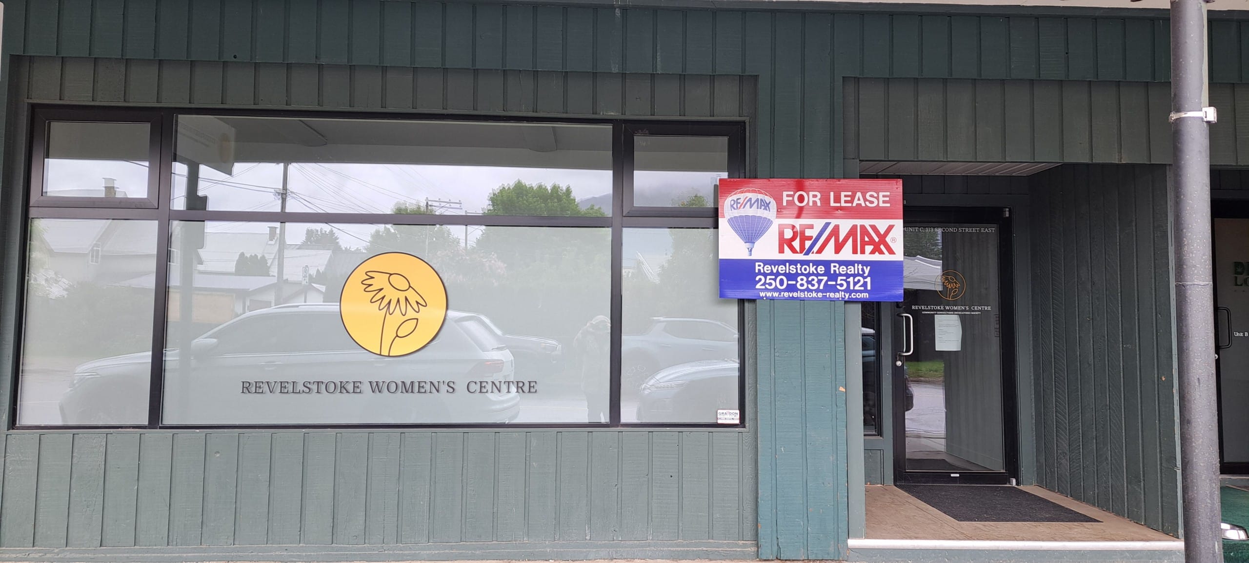 Front entrance of the Revelstoke Women's Centre with a re/max for sale sign posted beside a large window with the women's centre logo still in it.