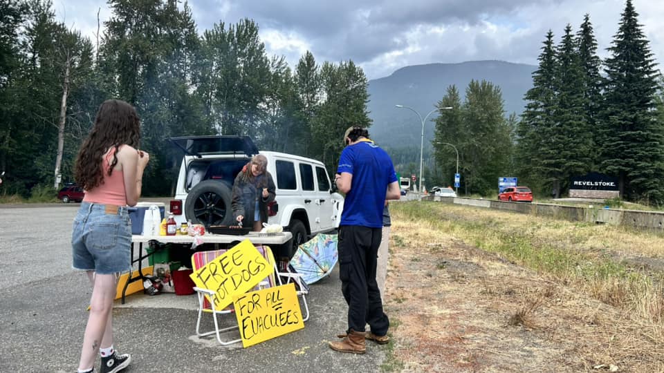 Two figures stand beside a makeshift hotdog stand along a busy highway. There is a grill on a plastic table and signs offering free hotdogs to evacuees.