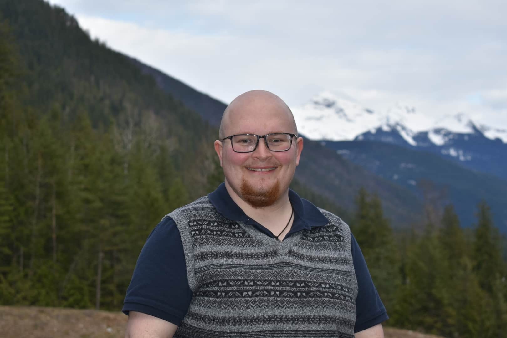 Revelstoke Mountaineer's community journalist Lys Morton, a white man with a shaved head and a small brown beard stands in front of a mountain range and smiles at the camera.