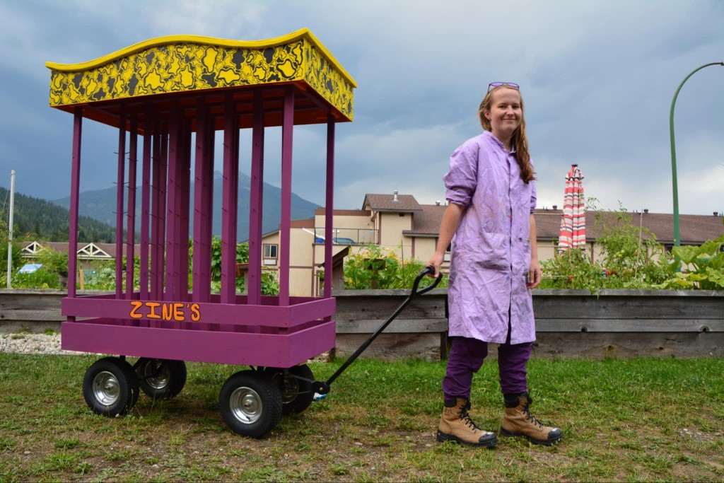 A purple and yellow wooden structure on top of a red wagon with ZINE written on top. A woman in a purple dress and leggings sands to the right of it with a smile on her face.