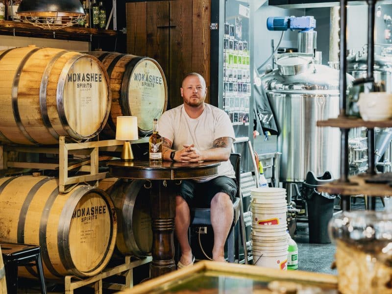 Monashee Spirits founder and distiller Josh McLafferty, a man in a white t-shirt with buzzed hair staring at the camera. He sits at a table in front of a wall of whiskey barrels.