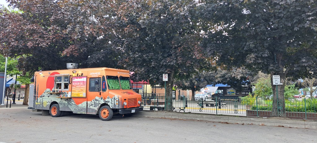 Twilight Bite food truck, an orange and green box truck, parked in front of Revelstoke's Grizzly Plaza. The plaza gazebo is in the background behind some trees.