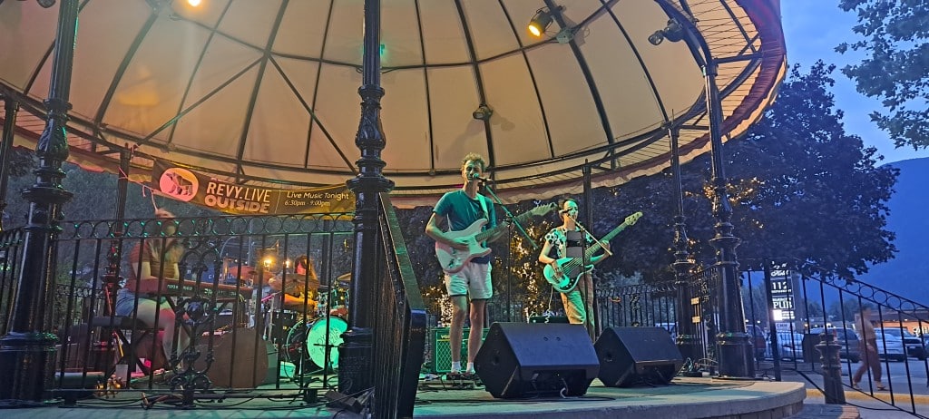 Two guitarists and a percussionist perform on a gazebo stage with green light backlighting them. A banner behind them promotes REVY.Live Outside