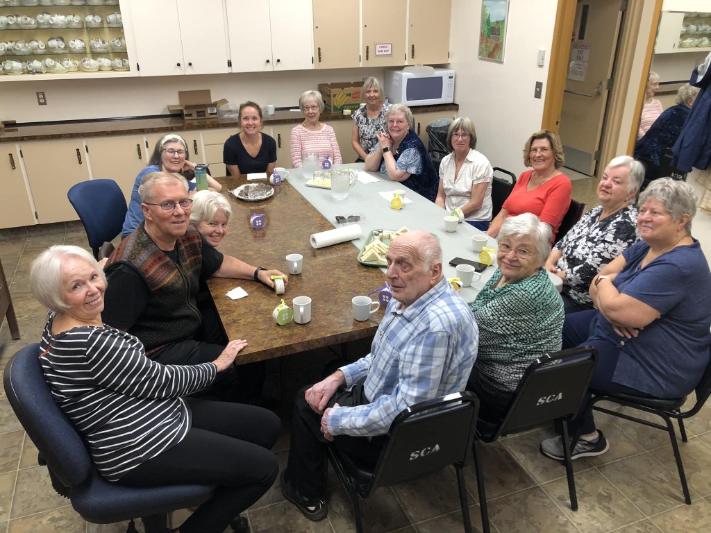 14 people sit around two tables in a community hall kitchen. They are all looking up at a camera and smiling, some leaning back or forth to be seen around the person sitting in front of them.