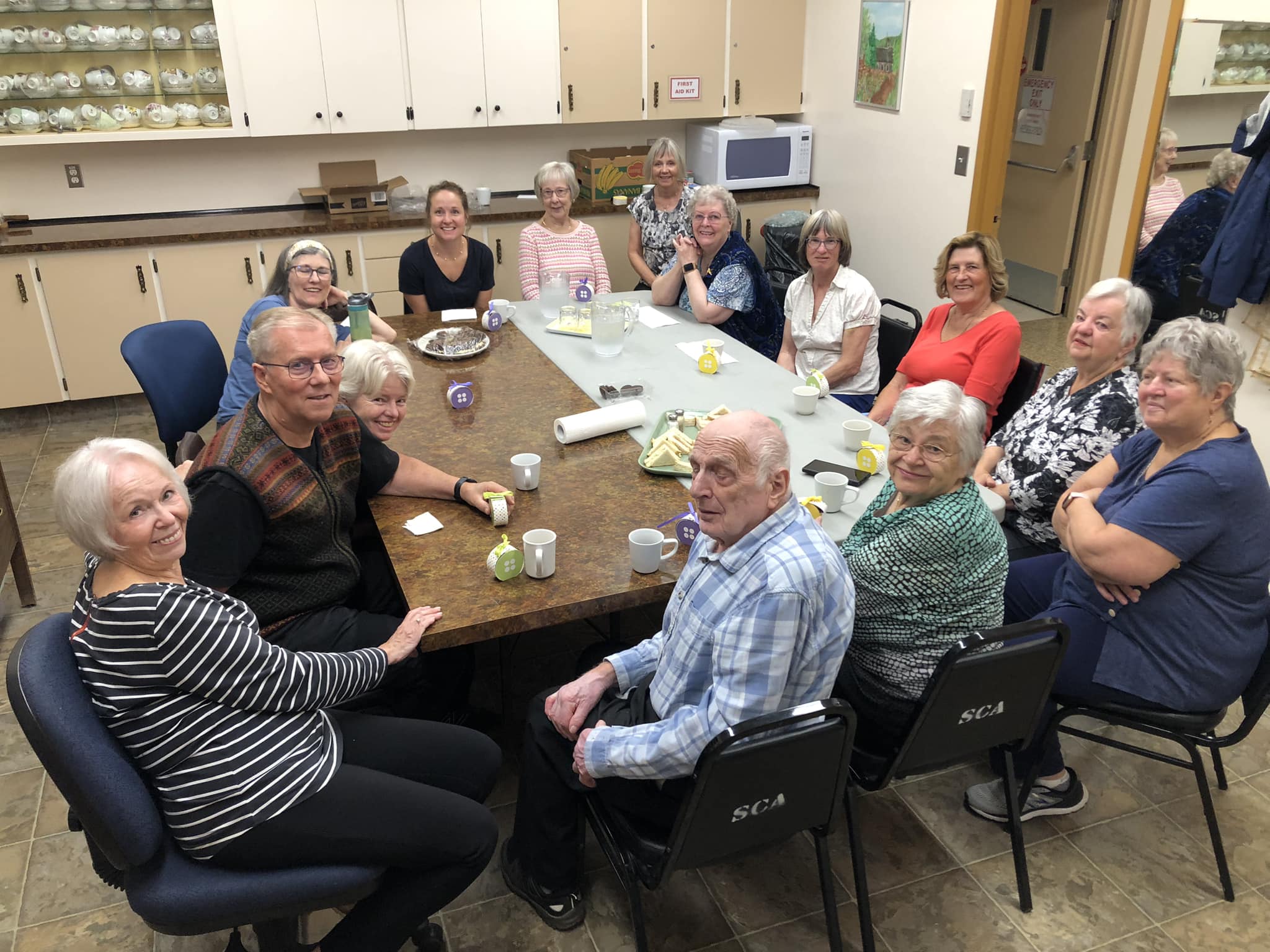14 people sit around two tables in a community hall kitchen. They are all looking up at a camera and smiling, some leaning back or forth to be seen around the person sitting in front of them.