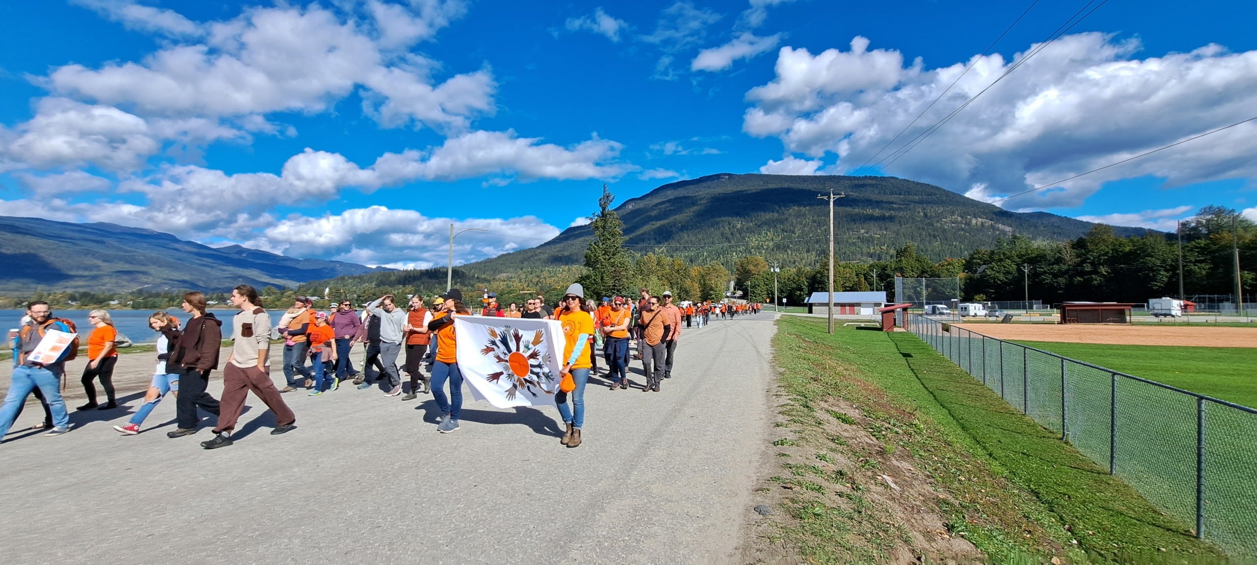 Orange Shirt Day 2024 participants marching down the road to Centennial Park in Revelstoke. There are two leaders holding a banner of an orange sun surrounded by hand prints.